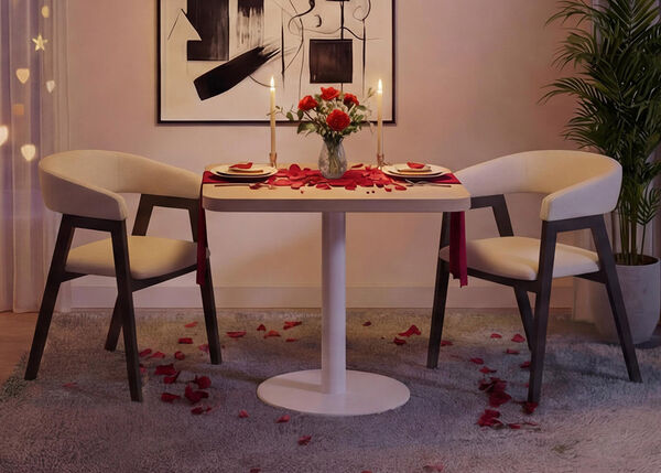 Allestimento romantico per la cena in un soggiorno moderno con un piccolo tavolo rotondo bianco e due sedie imbottite, apparecchiato con piatti, candele, un vaso di rose rosse e petali di rosa sparsi sul tavolo e sul tappeto, incorniciato da morbide luci a tenda, arte astratta alle pareti e una pianta verde da interno per un&rsquo;atmosfera accogliente di San Valentino.