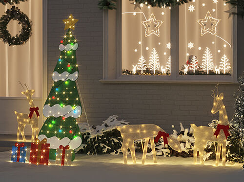 Decorazione natalizia esterna con figure di renne illuminate con fiocchi rossi, un albero verde e bianco illuminato con luci colorate e regali incartati sulla neve, davanti a una casa con decorazioni luminose di stelle e alberi alle finestre.