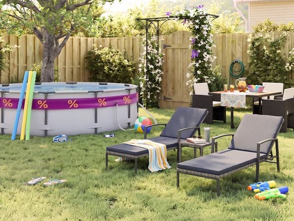 Giardino sul retro soleggiato con piscina fuori terra, tubi galleggianti, occhialini da nuoto, pallone da spiaggia e infradito sull&rsquo;erba. Due lettini prendisole in metallo nero con cuscini grigi sono sistemati accanto a un tavolino, mentre un set da pranzo da esterno con sedie scure intrecciate si trova vicino a un pergolato fiorito, una staccionata in legno e piante da giardino.