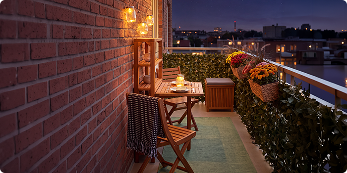Piccolo balcone con sedie e tavolo in legno, illuminato da lanterne e candele, decorato con fiori in vaso e verde, con vista sullo skyline della città di notte.
