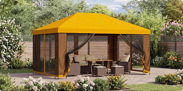 Gazebo da giardino giallo con tende a rete che ripara un salottino con tavolo su un patio, circondato da piante fiorite in un giardino soleggiato.