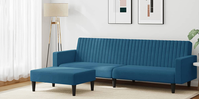 Un moderno divano componibile in velluto blu con trapuntatura a canali verticali e pouf coordinato. Il divano ha gambe in legno nero ed è posizionato in un soggiorno luminoso, con quadri incorniciati alla parete e una lampada da terra nell’angolo.